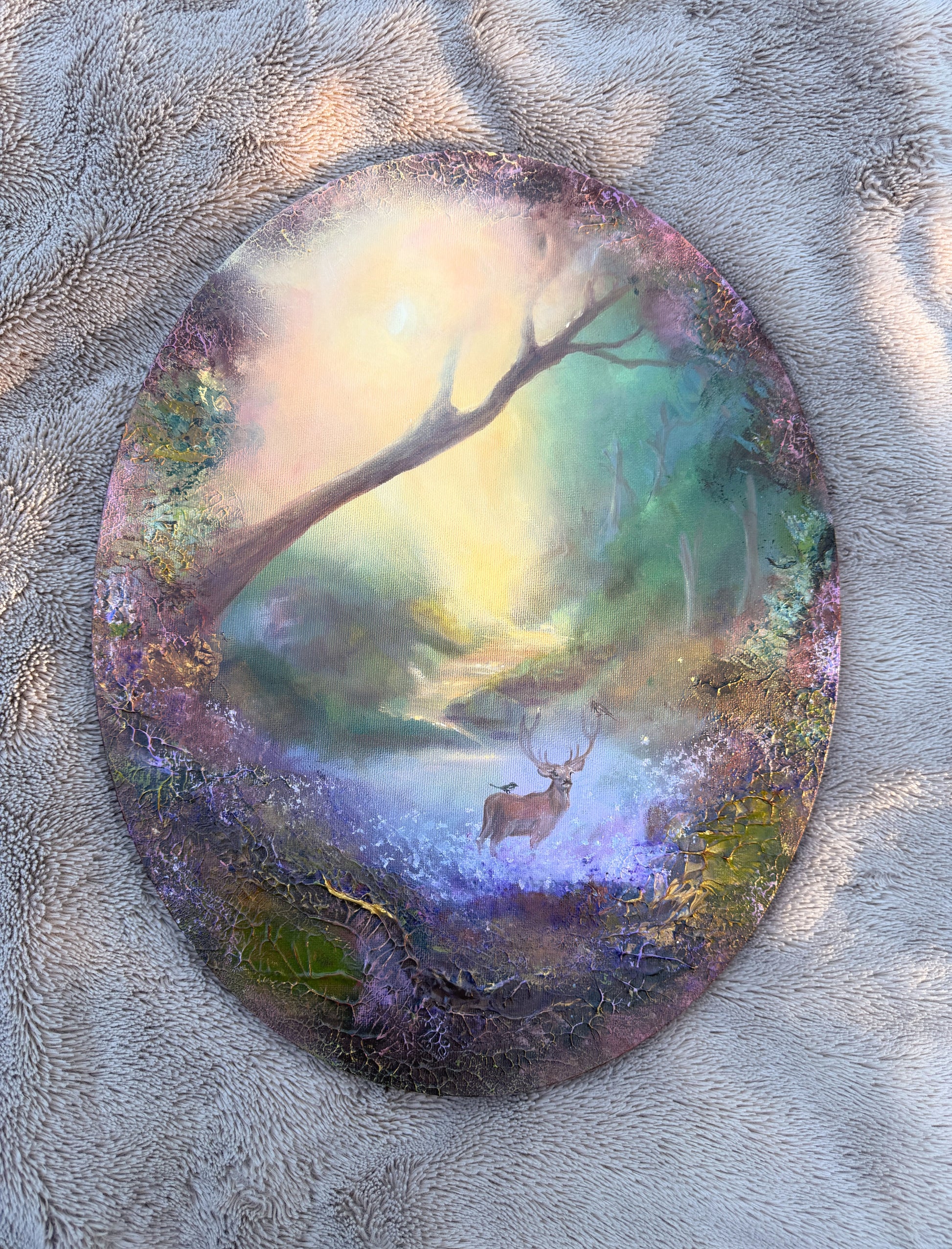An oval acrylic art print depicting a mystical forest scene with a stag under a glowing misty sky, displayed on a textured gray blanket.