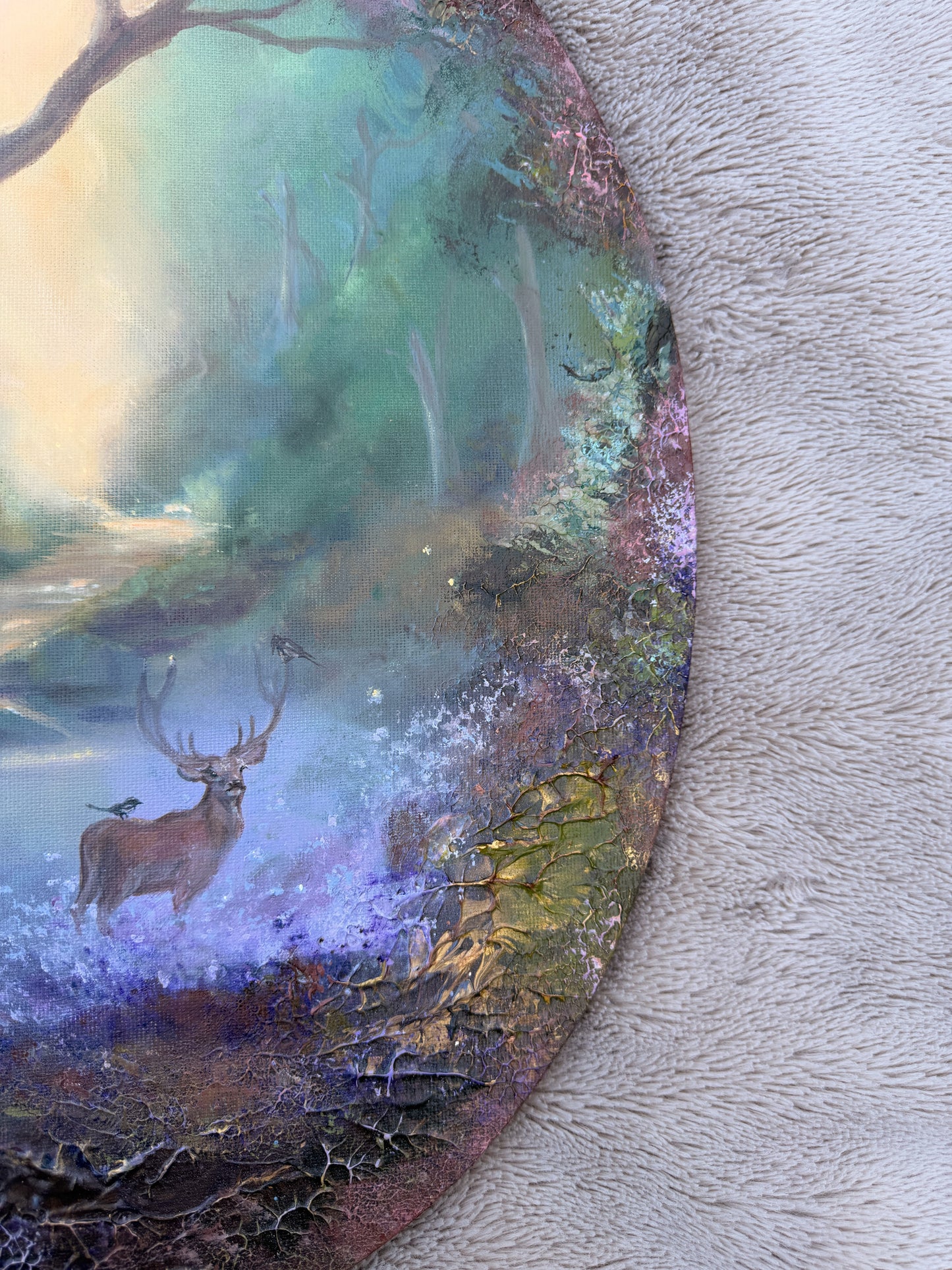 A close-up of a textured circular painting depicting a deer in a misty forest with purple, green, and gold tones, set against a light beige textured background.