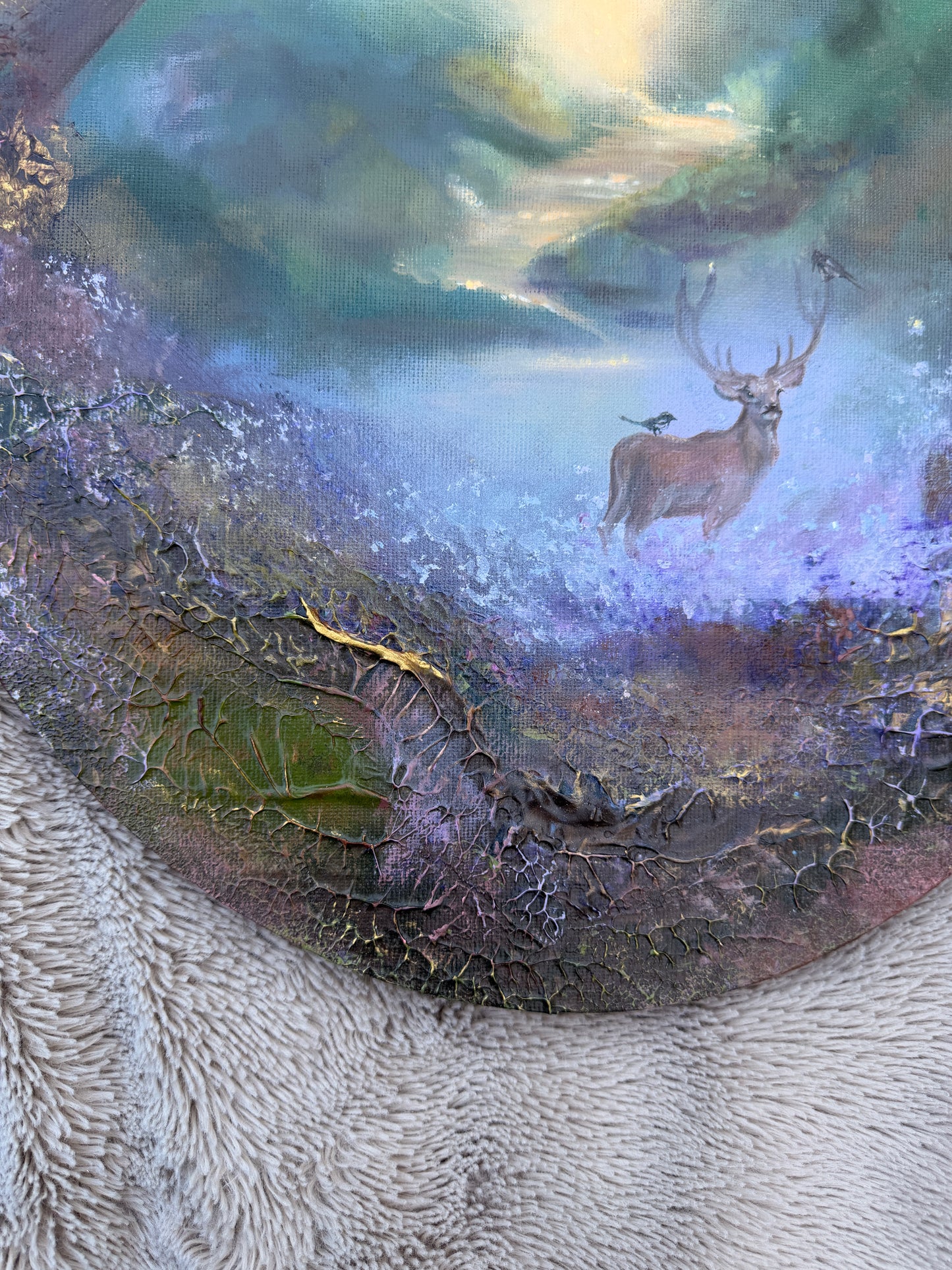 A close-up of a textured circular artwork depicting a stag in a misty forest with birds, layered over a cracked earthy surface and resting on a white textured fabric.