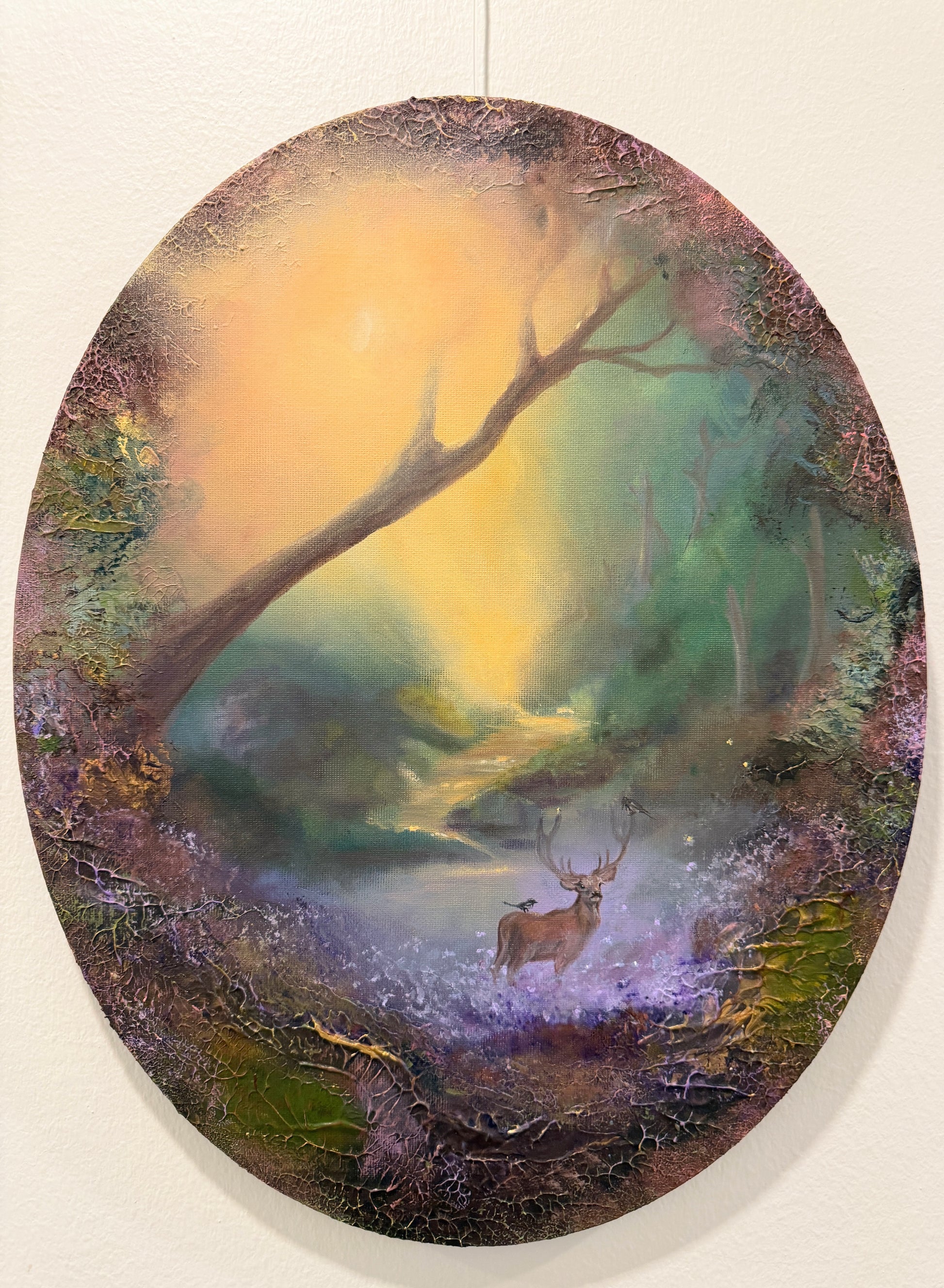 An oval textured painting depicting a mystical forest scene with a deer, hanging on a light beige wall.